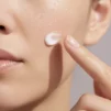 Close-up of a woman's cheek with water droplets and a dab of moisturiser on her fingertip
