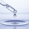 Glass dropper releasing a clear drop of hyaluronic acid serum above water ripples