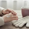 Hands applying hand cream beside a knitted glove and a cup of tea
