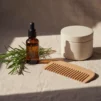 Unlabelled dropper bottle of hair oil with fresh rosemary sprigs and a wooden comb on cream linen