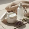 Unlabelled jar of gentle cream, pump bottle and dropper bottle on a cream linen cloth beside a soft towel